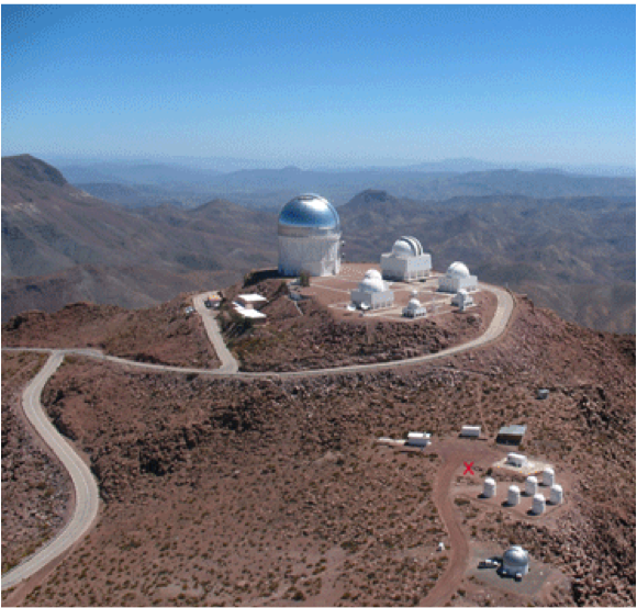 PROMPT is a cluster of small telescope domes situated just down slope from several large telescopes on the top of the mountain. There is blue sky above and distant mountains in the background.  One can also see the a white road surrounding the telescopes on the mountain top and a rougher road leading to PROMPT and other small domes near PROMPT.
